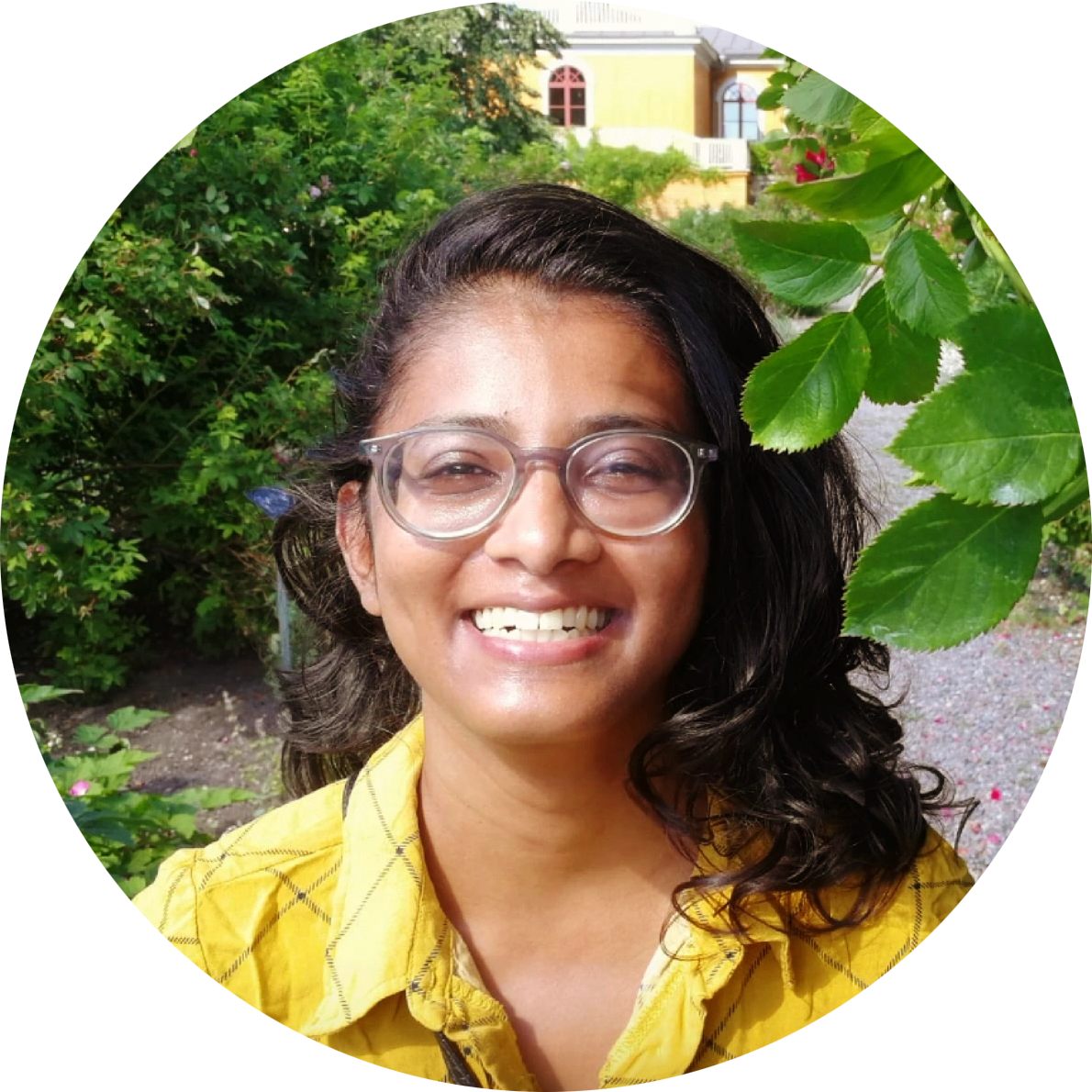 Woman of Indian descent wearing rounded glasses and a yellow shirt smiling broadly.